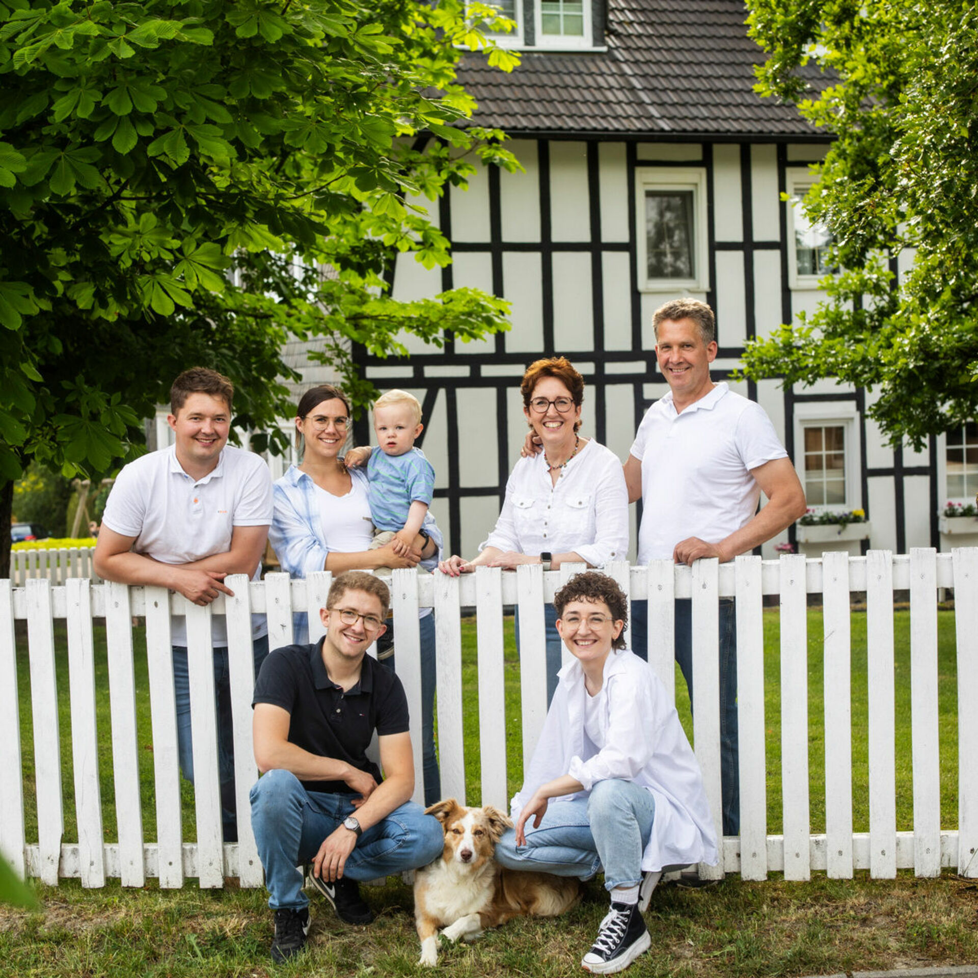 Personen vor unter hinter Zaun vor einem Fachwerkhaus mit Hund