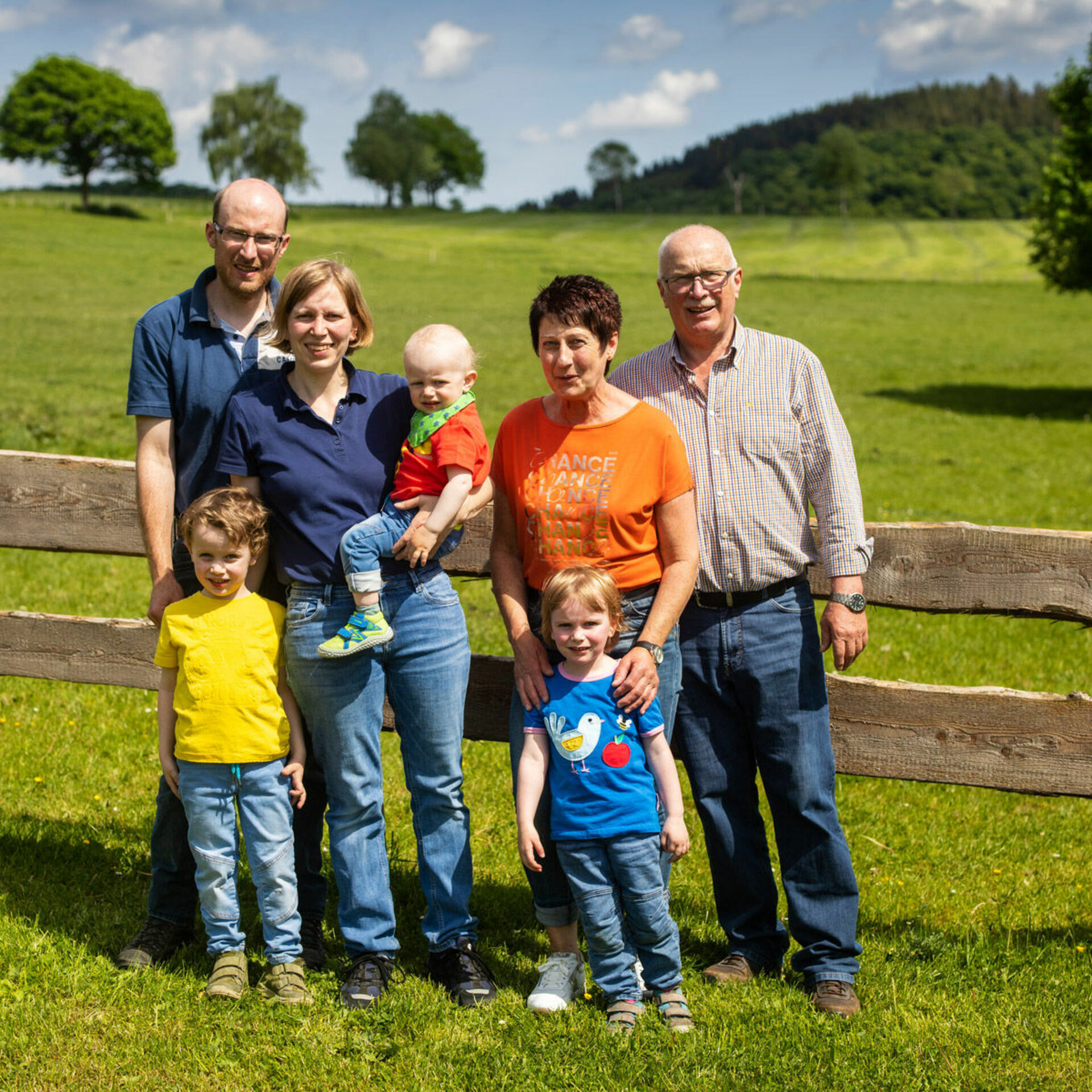 Gastgeberfamilie, bestehend aus 4 Erwachsenen und 4 Kindern,  stehend auf Wiese vor Zaun. 